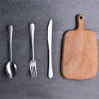 Wooden cutting board with silver spoon, fork, and knife on a gray surface