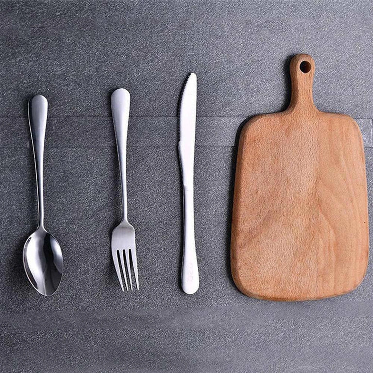 Wooden cutting board with silver spoon, fork, and knife on a gray surface