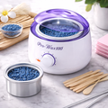 White wax warmer with purple lid on a marble surface, surrounded by blue wax beads and wooden sticks.